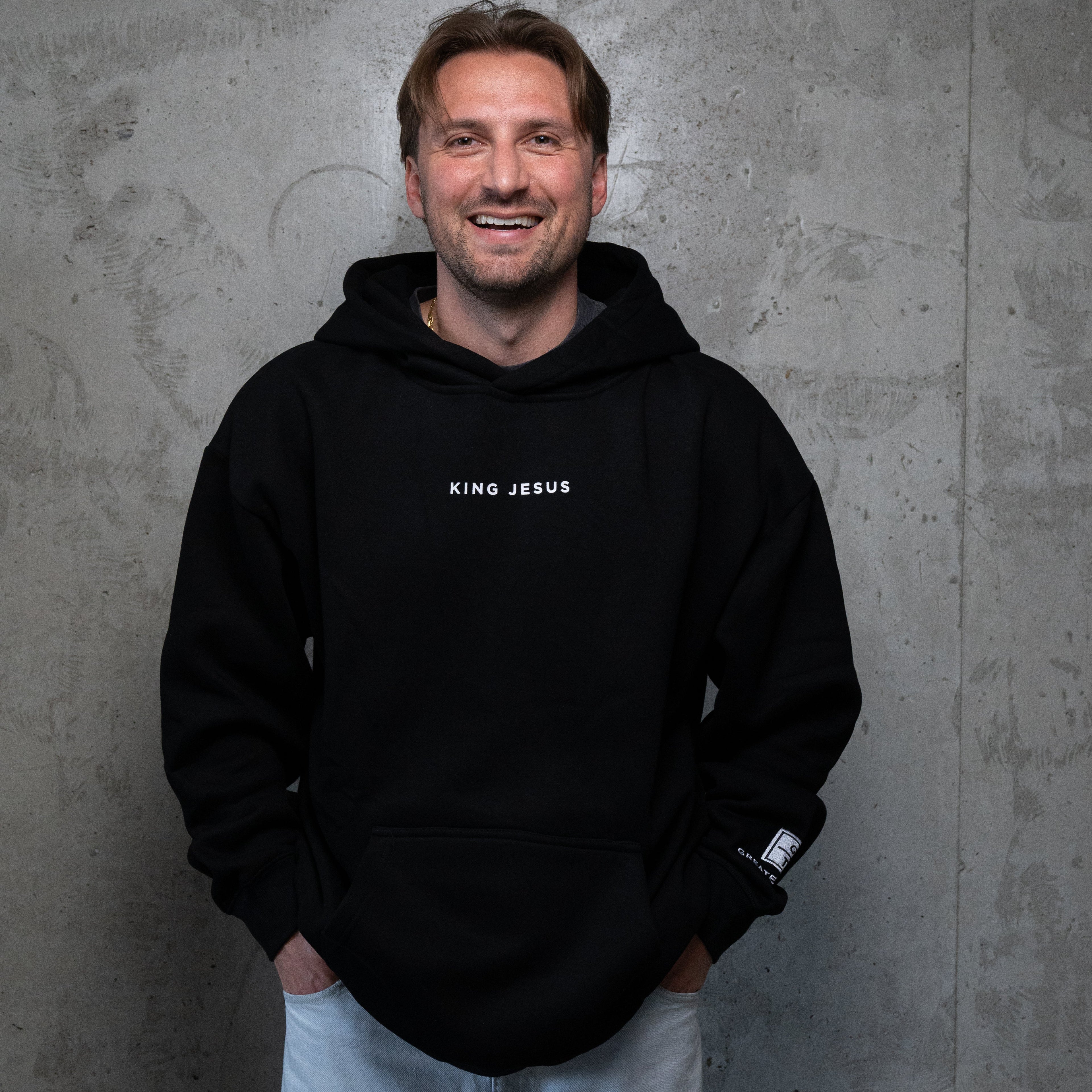 A smiling man wearing a black pullover hoodie with 'KING JESUS' printed in white on the chest, paired with light gray pants, standing against a textured gray concrete wall.