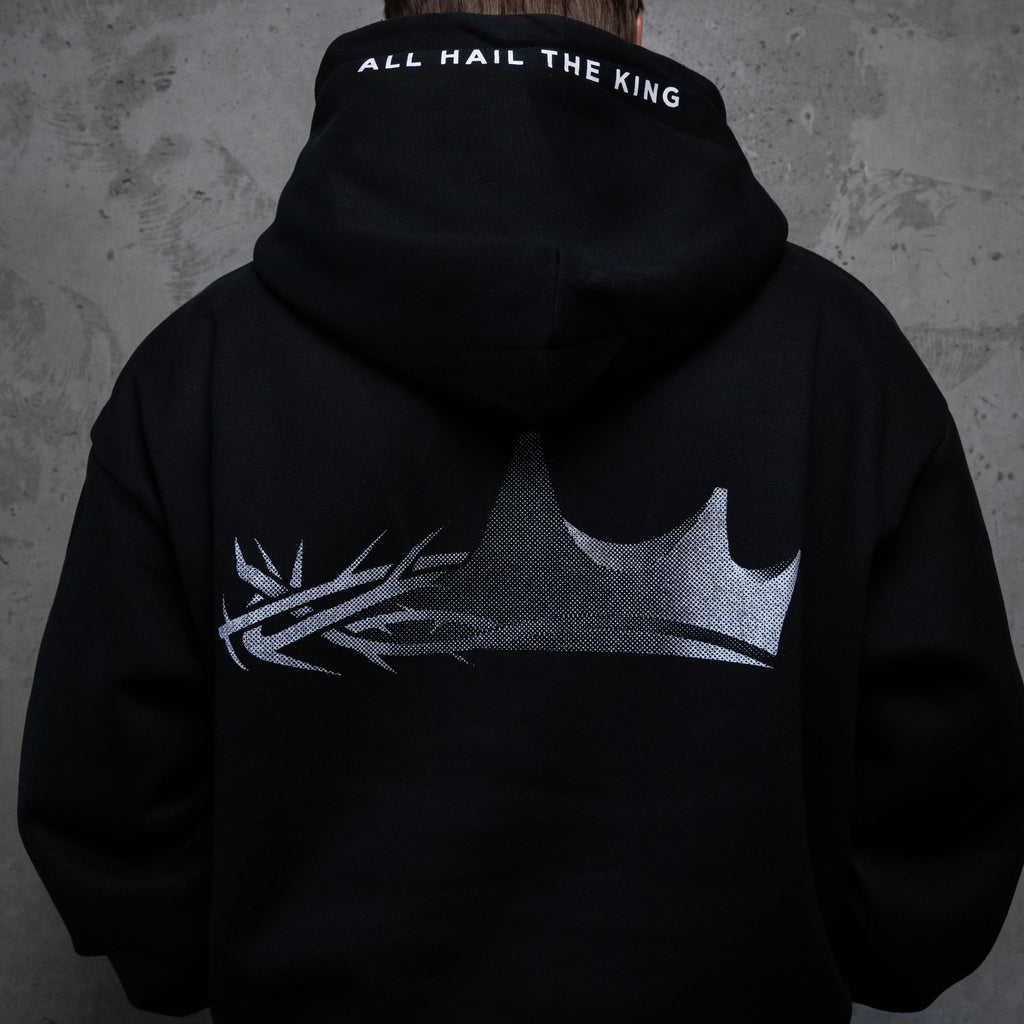 A person viewed from behind wearing a black hoodie with a large silver crown graphic and the words 'ALL HAIL THE KING' printed on the inside of the hood, standing against a textured gray wall.