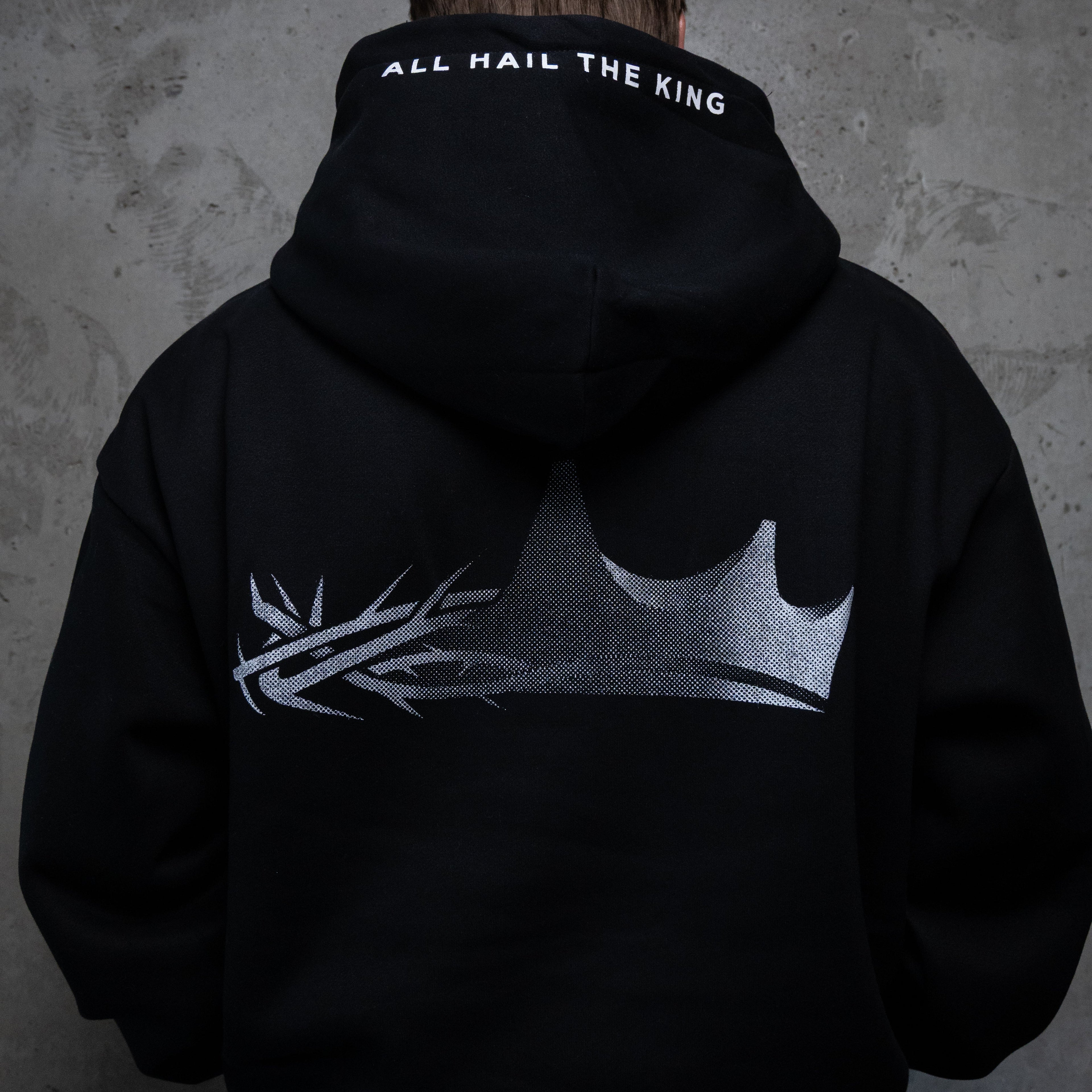 A person viewed from behind wearing a black hoodie with a large silver crown graphic and the words 'ALL HAIL THE KING' printed on the inside of the hood, standing against a textured gray wall.