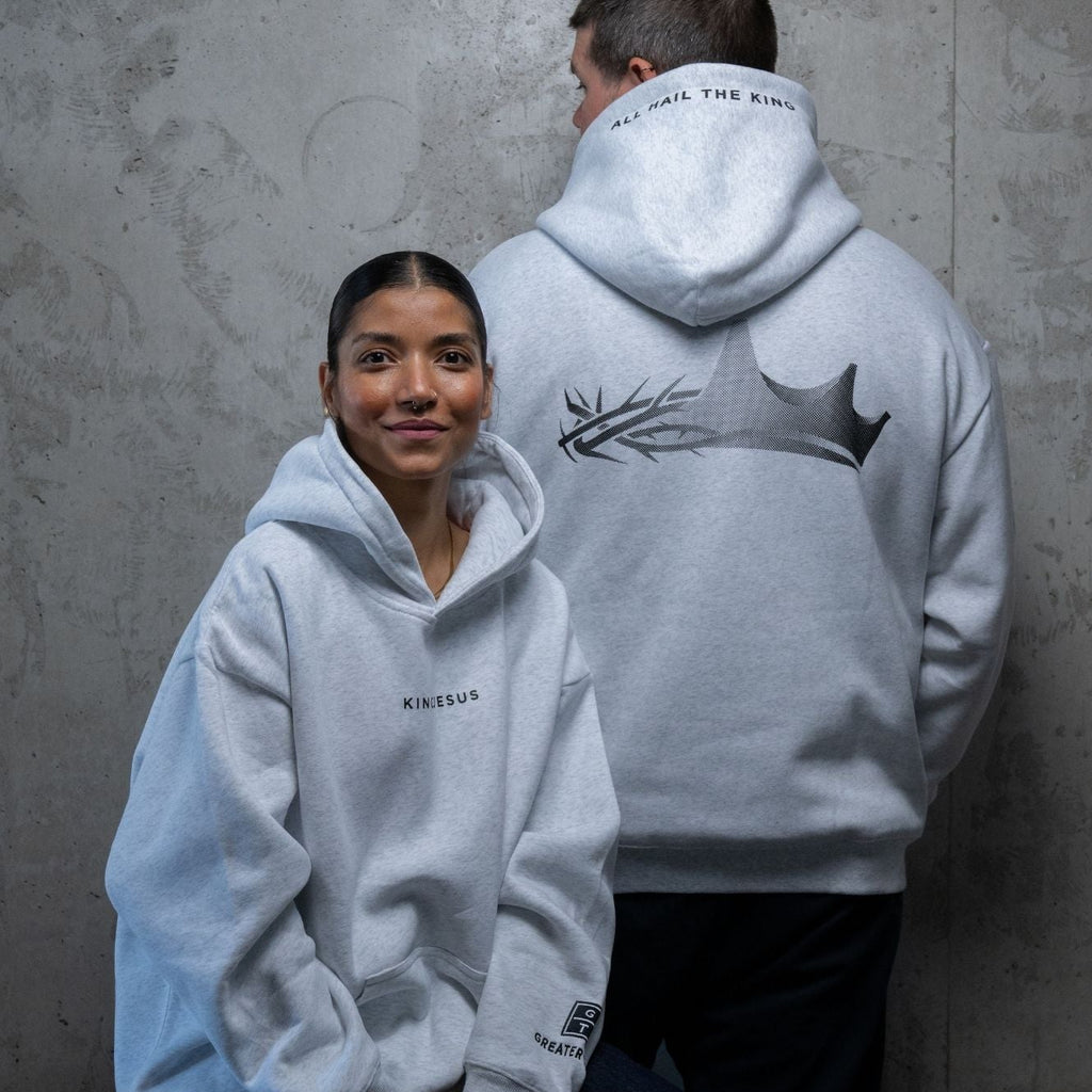 Two models wearing light gray hoodies from the back and front, standing against a textured gray concrete wall. The model in the back wears a large black graphic of a crown and branch on the back, while the model in the front wears a small black logo on the left chest.
