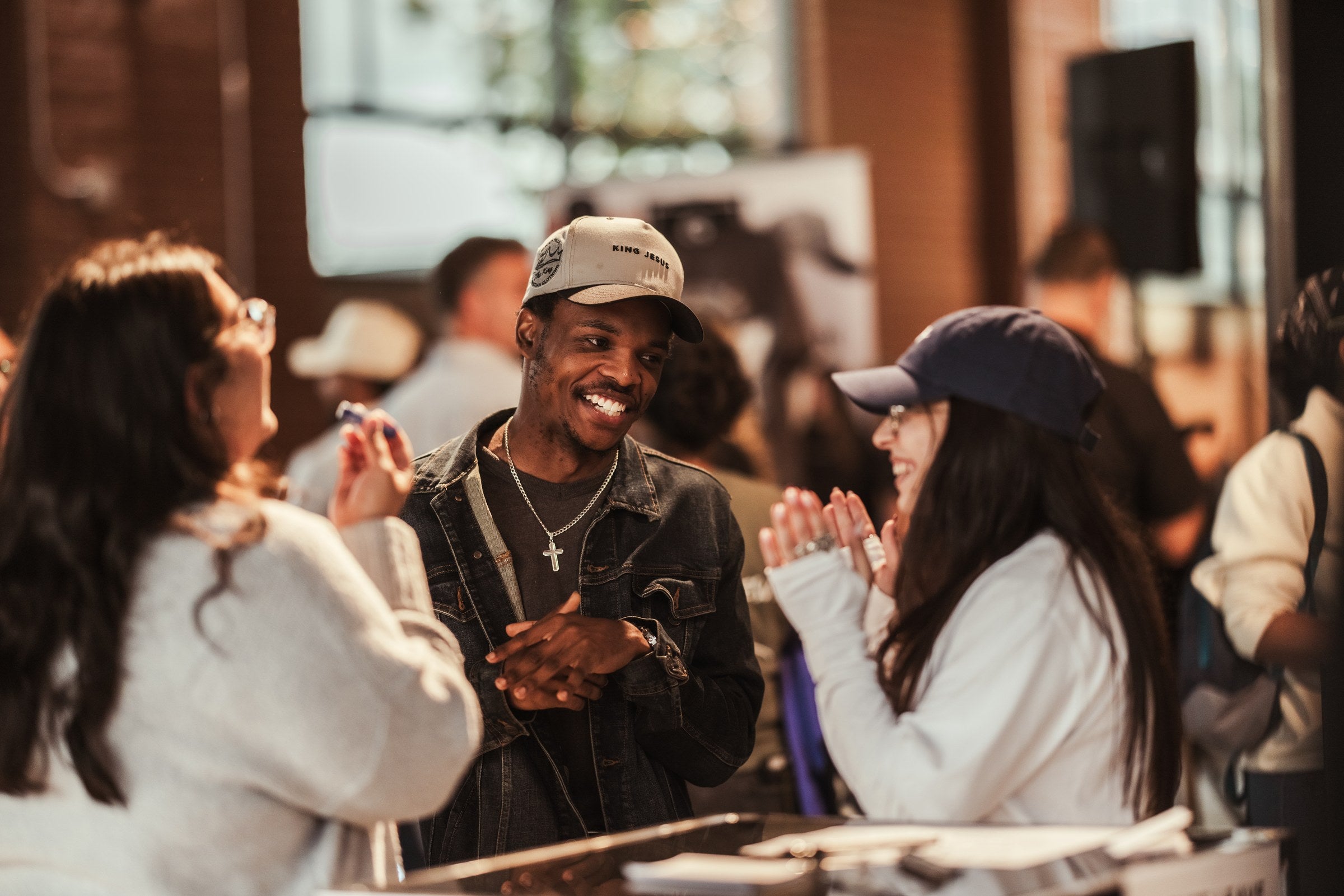 Three people talking and laughing together before a GREATER THAN worship night begins, one wearing a KING JESUS cap.
