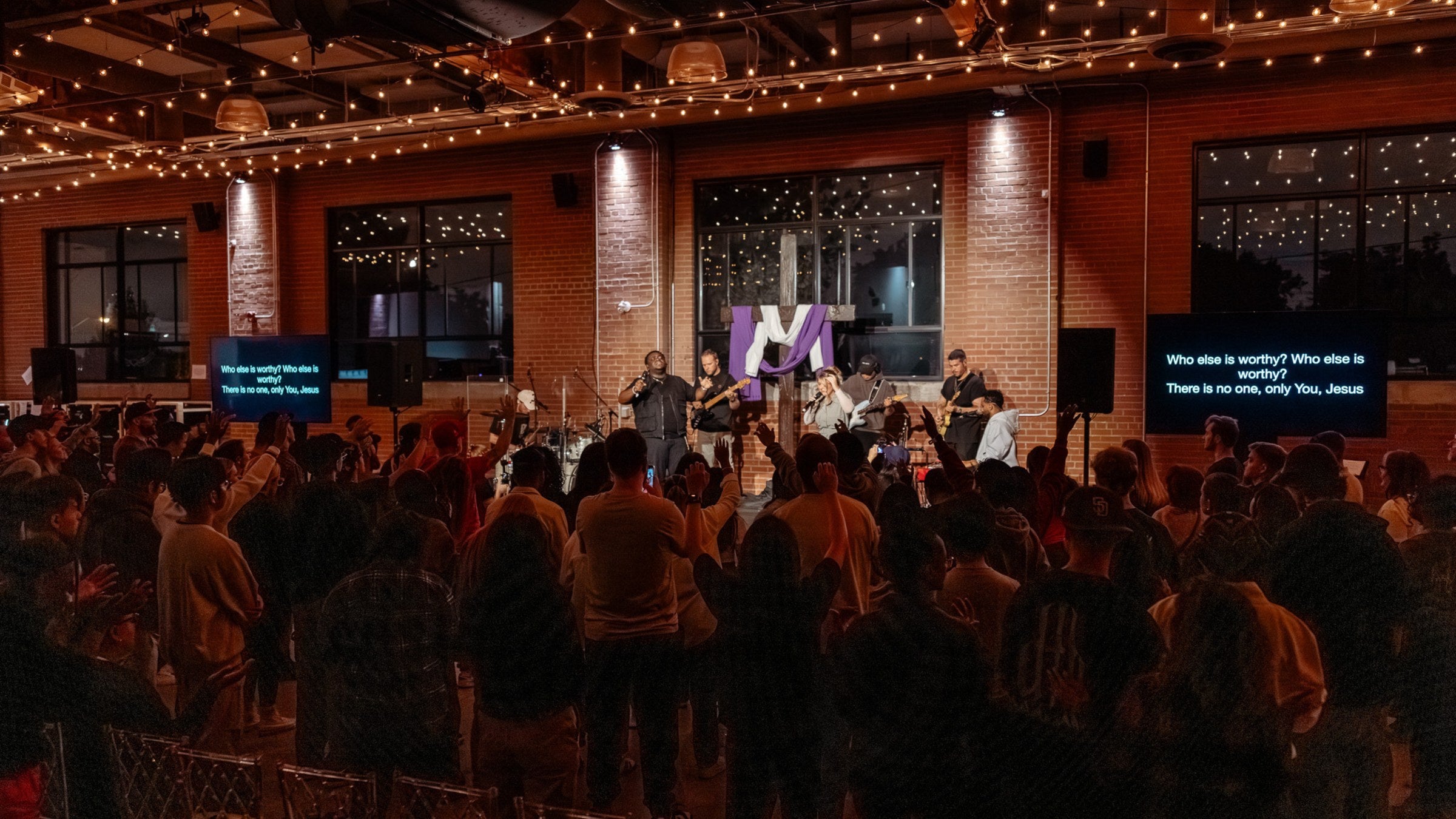 A full room of people standing with hands raised at a GREATER THAN worship night.