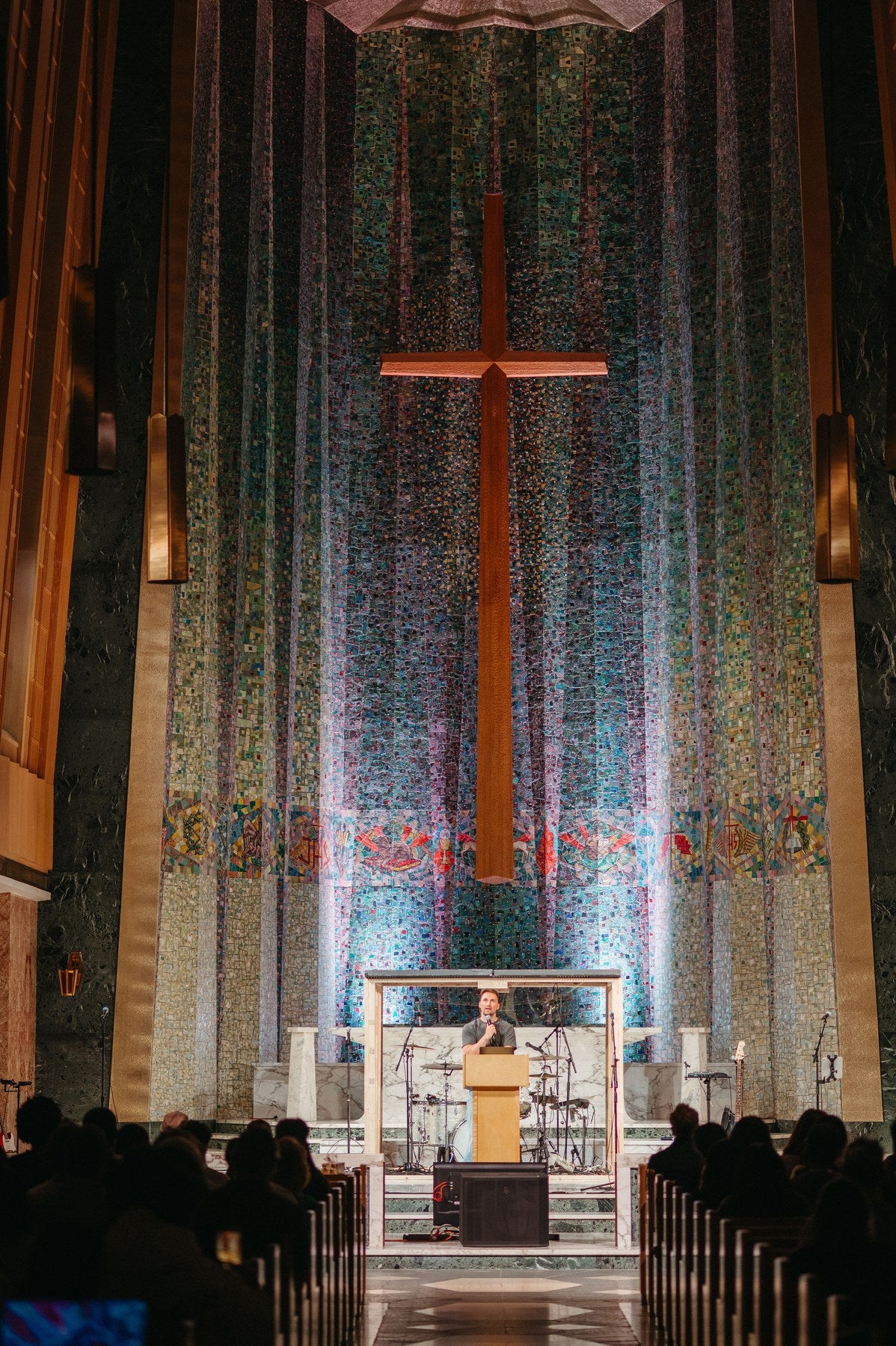 Pastor Ty Smith preaching from a wooden podium in Tyndale University Chapel, beneath a tall wooden cross set against a floor-to-ceiling mosaic wall.