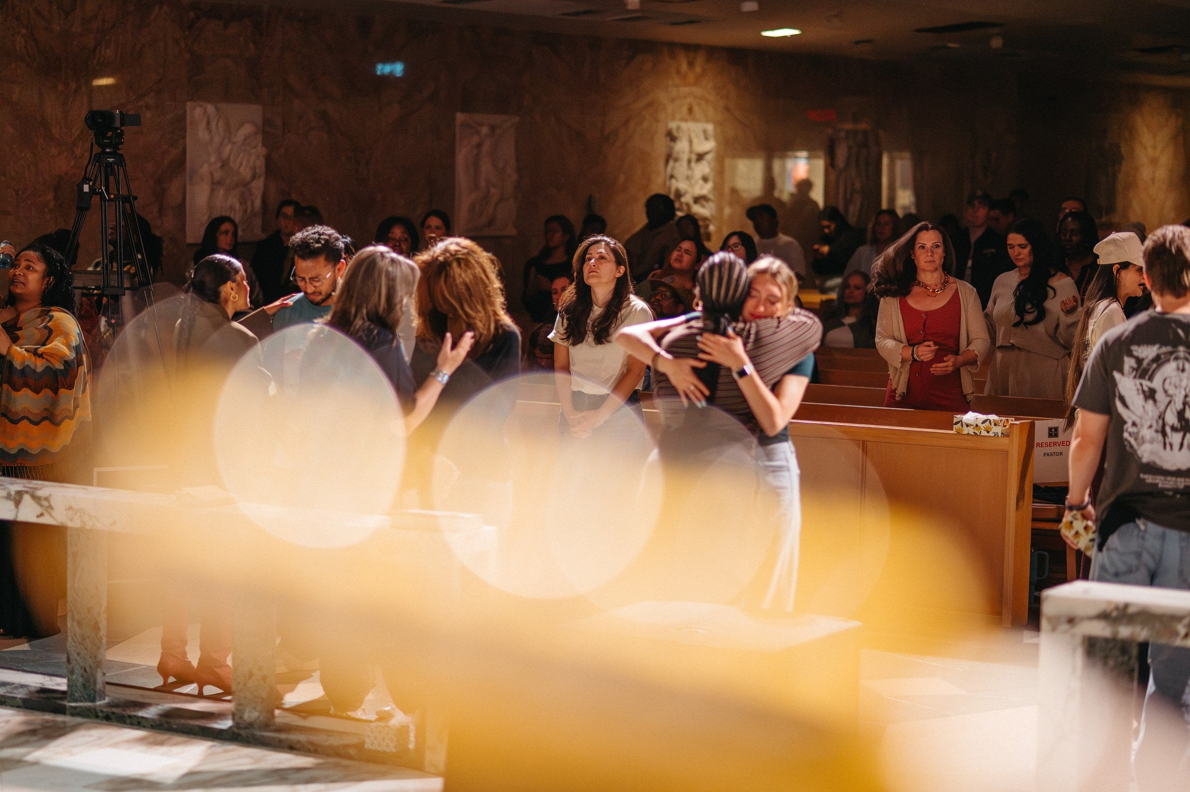 People gathered at the front of the chapel during the prayer pit, praying together and embracing, while others stand watching from the pews.