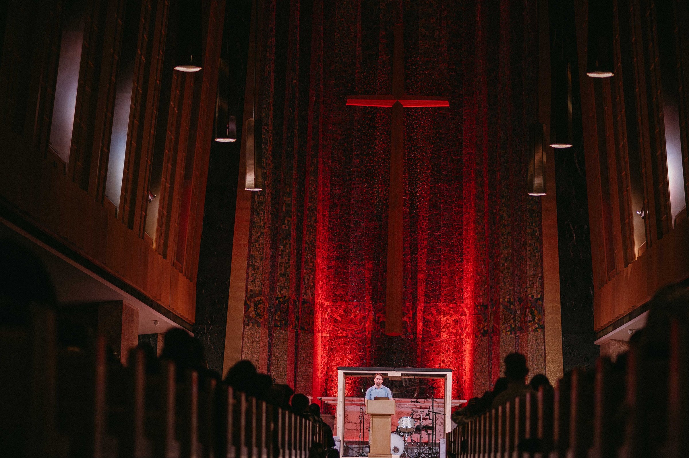 The congregation seated in Tyndale University Chapel during a GREATER THAN worship night, facing a tall wooden cross lit in deep red against the chapel's full-height mosaic wall.