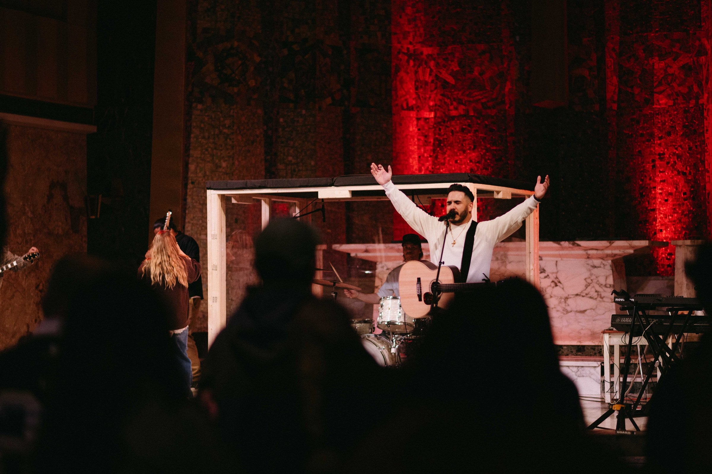 A GREATER THAN worship leader singing with both arms raised at the front of the chapel, with the band and drummer behind him under red stage lighting.