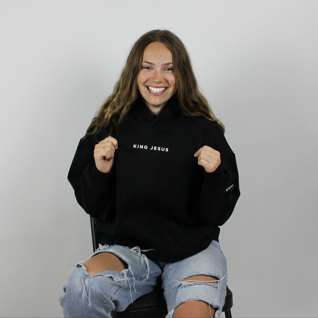 A person wearing an oversized black hoodie with the text 'King Jesus' on it, sitting on a chair with a neutral background.