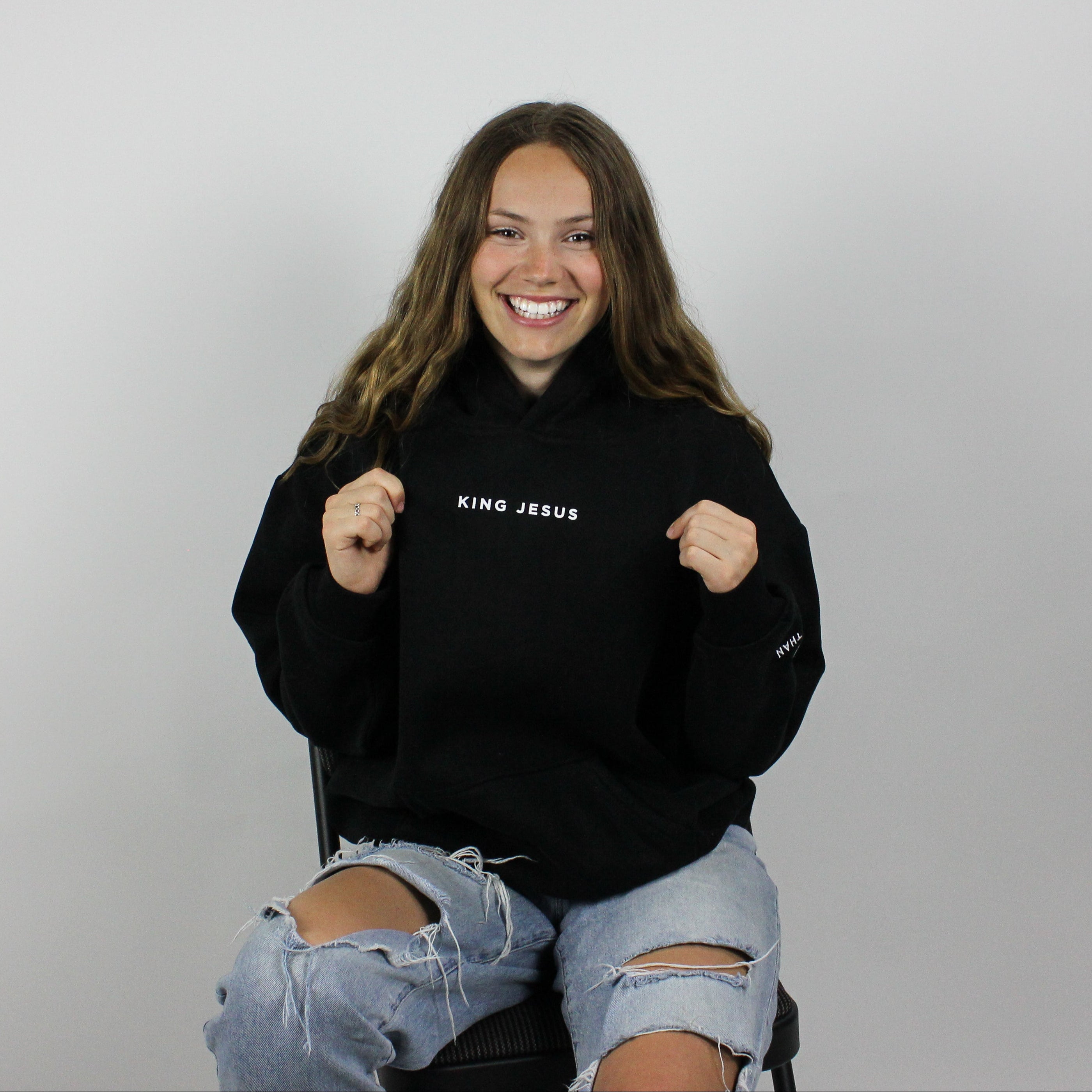 A person wearing an oversized black hoodie with the text 'King Jesus' on it, sitting on a chair with a neutral background.